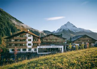 Ein gemütliches Hotel im alpinen Stil, umgeben von grünen Wiesen und Bergen. Im Hintergrund ist ein schneebedeckter Gipfel zu sehen.