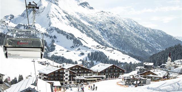Eine malerische Berglandschaft mit schneebedeckten Gipfeln und einer Skiliftbahn. Im Vordergrund sind gemütliche Chalets und Skifahrer auf der Piste zu sehen.