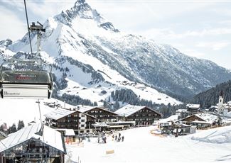 Eine malerische Berglandschaft mit schneebedeckten Gipfeln und einer Skiliftbahn. Im Vordergrund sind gemütliche Chalets und Skifahrer auf der Piste zu sehen.