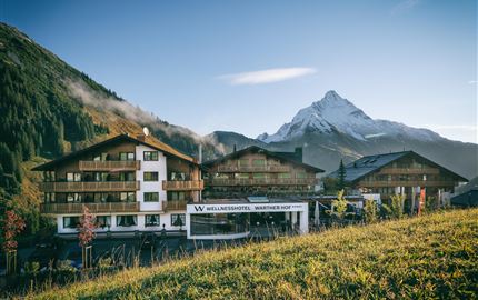 Ein gemütliches Hotel im alpinen Stil, umgeben von grünen Wiesen und Bergen. Im Hintergrund ist ein schneebedeckter Gipfel zu sehen.