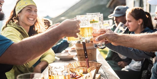Eine fröhliche Gruppe von Menschen stößt mit Biergläsern an. Im Hintergrund ist eine malerische Berglandschaft zu sehen.