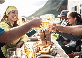 Eine fröhliche Gruppe von Menschen stößt mit Biergläsern an. Im Hintergrund ist eine malerische Berglandschaft zu sehen.