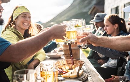 Eine fröhliche Gruppe von Menschen stößt mit Biergläsern an. Im Hintergrund ist eine malerische Berglandschaft zu sehen.