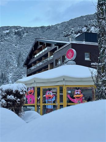 Ein gemütliches Restaurant mit großen Fenstern, das mit bunten Figuren dekoriert ist. Die Umgebung ist schneebedeckt und von Bergen umgeben.
