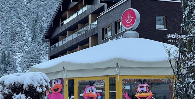 Ein gemütliches Restaurant mit großen Fenstern, das mit bunten Figuren dekoriert ist. Die Umgebung ist schneebedeckt und von Bergen umgeben.