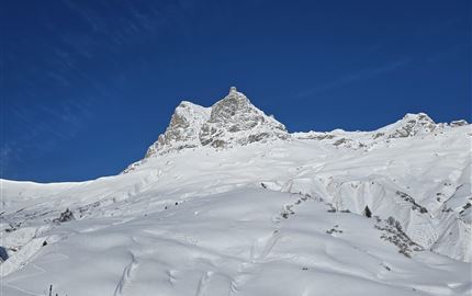 Eine schneebedeckte Landschaft mit Bergen und klarem blauen Himmel. Im Vordergrund sind mehrere Gebäude in einer ruhigen Umgebung zu sehen.