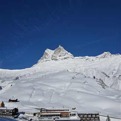 A snow-covered landscape with mountains and a clear blue sky. In the foreground, several buildings can be seen in a quiet environment.