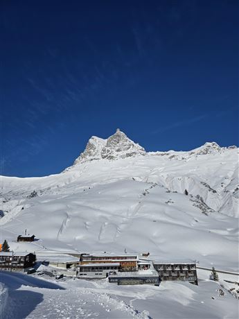 A snow-covered landscape with mountains and a clear blue sky. In the foreground, several buildings can be seen in a quiet environment.