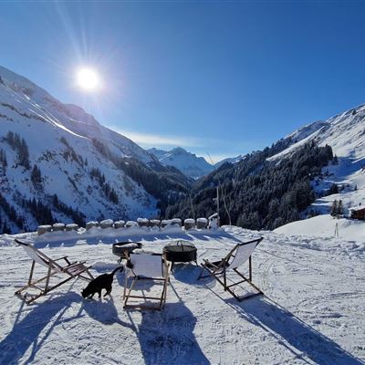 Eine winterliche Berglandschaft mit schneebedeckten Hügeln und strahlend blauem Himmel. Zwei Liegestühle und ein Hund stehen im Vordergrund.