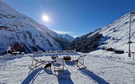 Eine winterliche Berglandschaft mit schneebedeckten Hügeln und strahlend blauem Himmel. Zwei Liegestühle und ein Hund stehen im Vordergrund.