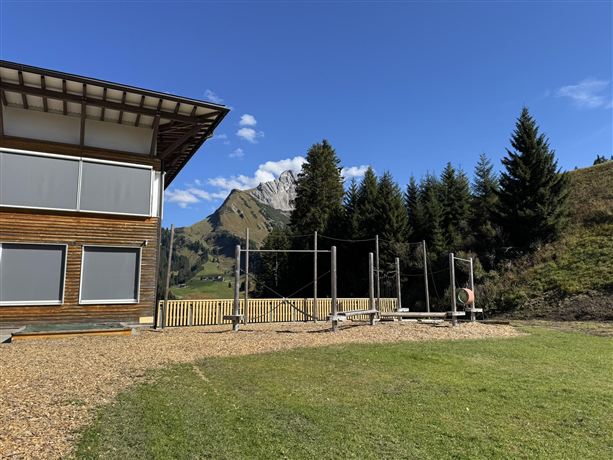 Ein modernes Gebäude in einer grünen Wiese mit Blick auf Berge. Umgeben von Nadelbäumen und klarem Himmel.