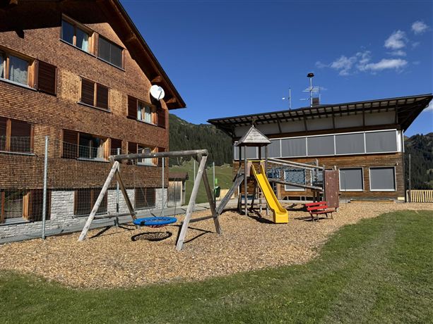 Ein Spielplatz mit Schaukeln und einem Spielturm in einer ländlichen Umgebung. Im Hintergrund sind zwei Gebäude und blauer Himmel zu sehen.
