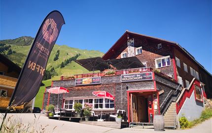 Ein rustikales Restaurant in den Bergen mit einer terrassenartigen Außensitzfläche. Die Umgebung ist grün und passt perfekt zur alpinen Atmosphäre.
