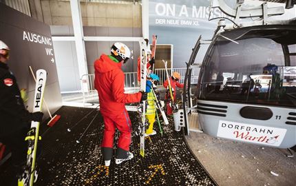 Eine Gruppe von Skifahrern wartet an einer Gondelbahn. Im Hintergrund ist die Gondel und der Ausgang sichtbar.