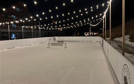 Eine beleuchtete Eisbahn bei Nacht mit festlichen Lichtern. Der Platz ist leer und umgeben von Schnee.