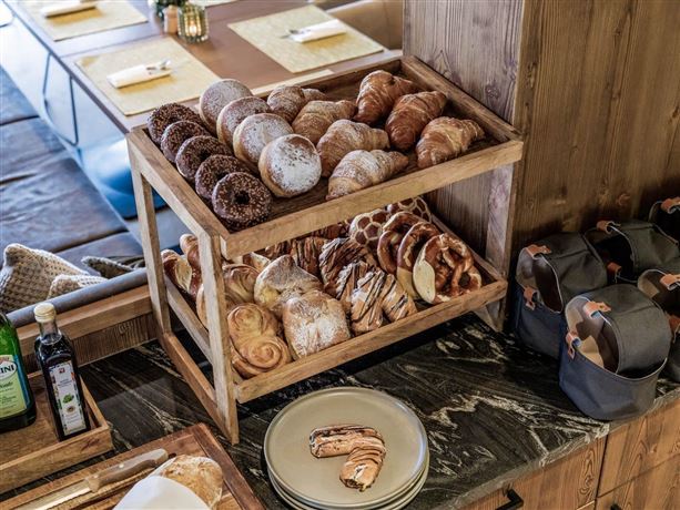 Ein Holzregal mit verschiedenen frisch gebackenen Brötchen und Croissants. Auf einem Tisch im Hintergrund sind gedeckte Plätze zu sehen.