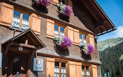 Ein charmantes Holzhaus mit bunten Blumenkästen an den Fenstern. Im Hintergrund sind grüne Berge und ein blauer Himmel zu sehen.