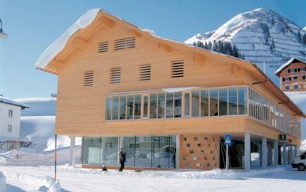 Ein modernes Holzgebäude in einer verschneiten Umgebung. Im Hintergrund sind Berge und eine klare blaue Himmel zu sehen.