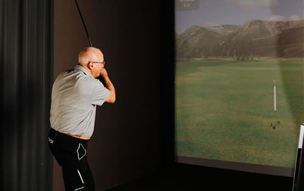 Ein Mann spielt Golf in einem Indoor-Simulator. Im Hintergrund ist eine virtuelle Golfbahn zu sehen.