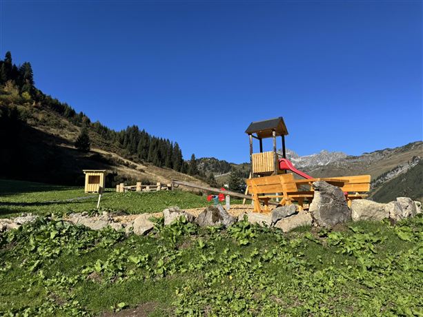 Ein Spielplatz mit einer Rutsche steht auf einer Wiese in den Bergen. Im Hintergrund sind hohe Berge und ein klarer blauer Himmel zu sehen.