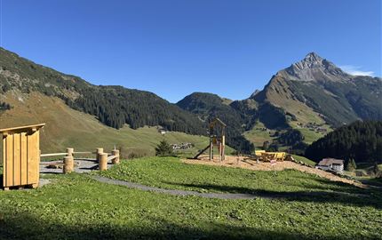 Eine malerische Berglandschaft mit grünen Wiesen und einem klaren blauen Himmel. Im Vordergrund sind Spielgeräte und eine Holzstruktur zu sehen.