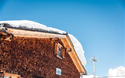 Ein traditionelles Holzhaus mit einem schneebedeckten Dach. Der klare Himmel im Hintergrund strahlt Sonnenlicht aus.