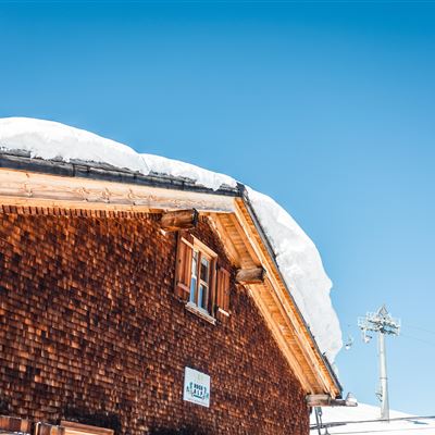Ein traditionelles Holzhaus mit einem schneebedeckten Dach. Der klare Himmel im Hintergrund strahlt Sonnenlicht aus.