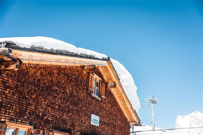Ein traditionelles Holzhaus mit einem schneebedeckten Dach. Der klare Himmel im Hintergrund strahlt Sonnenlicht aus.