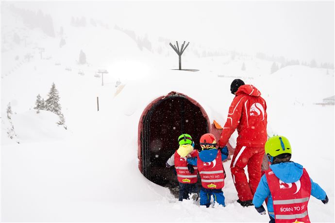 Ein Skilehrer bringt Kinder in einen schneebedeckten Bereich. Die Kinder tragen bunte Helme und Skijacken.