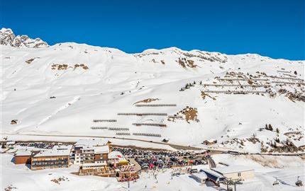 Eine schneebedeckte Berglandschaft mit einem klaren blauen Himmel. Im Vordergrund befindet sich ein Skilift und ein gemütliches Restaurant mit vielen Besuchern.