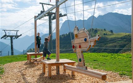A playground in the mountains with swings and a magnificent view of the peaks.  
Children are playing and enjoying the fresh air.