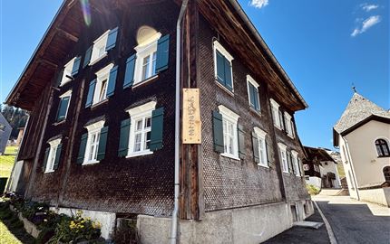 Ein traditionelles Schweizer Holzhaus mit dunkler Fassade und grünen Fensterläden. Die Umgebung ist von Grünflächen und blauen Himmel geprägt.