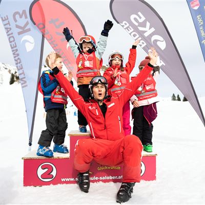 Eine Gruppe von Kindern in Skikleidung steht auf einem Siegertreppchen im Schnee. Ein Skilehrer freut sich mit ihnen und sie zeigen stolz ihre Medaillen.