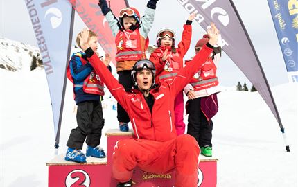 Eine Gruppe von Kindern in Skikleidung steht auf einem Siegertreppchen im Schnee. Ein Skilehrer freut sich mit ihnen und sie zeigen stolz ihre Medaillen.