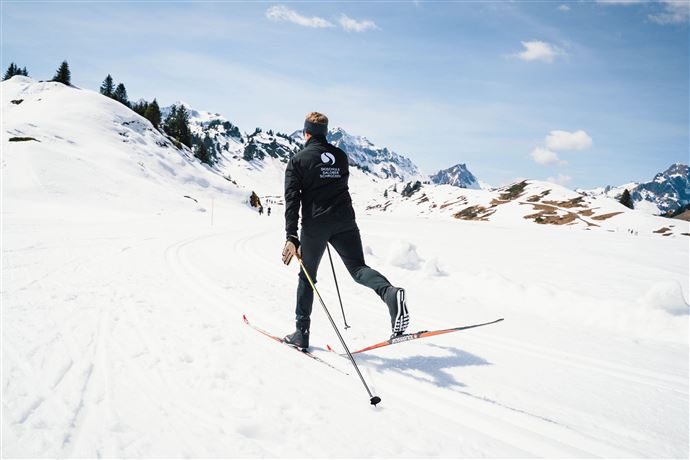 Eine Person läuft auf Langlaufskiern durch schneebedeckte Landschaften. Im Hintergrund sind verschneite Berge und ein klarer Himmel zu sehen.