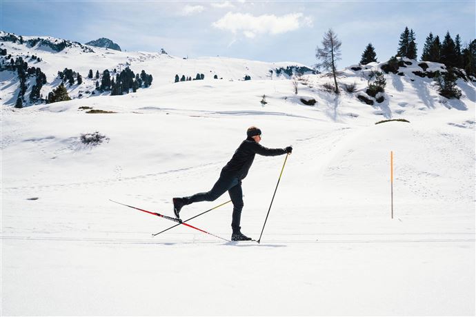 Eine Person macht Langlauf auf einer verschneiten Landschaft. Im Hintergrund sind Berge und Bäume sichtbar.