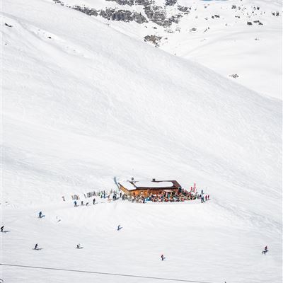 Eine verschneite Berglandschaft mit einer Almhütte und Skifahrern. Im Hintergrund sind schneebedeckte Gipfel und ein klarer blauer Himmel zu sehen.