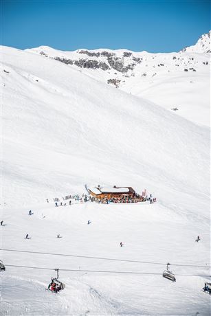 Eine verschneite Berglandschaft mit einer Almhütte und Skifahrern. Im Hintergrund sind schneebedeckte Gipfel und ein klarer blauer Himmel zu sehen.