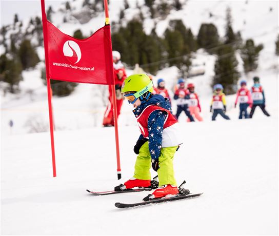 Ein kleiner Skifahrer mit grünem Helm übt neben einem roten Tor im Schnee. Im Hintergrund sind weitere Kinder in Skikursen zu sehen.