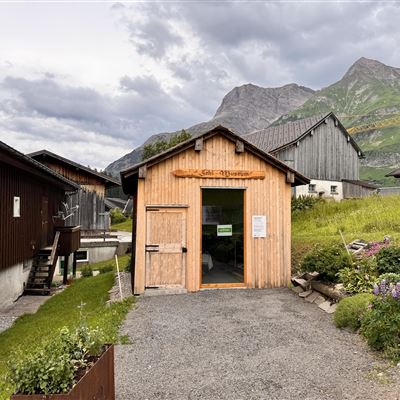 Ein Holzgebäude mit einer einladenden Eingangstür steht inmitten von grünen Wiesen und Bergen. Die Wolken am Himmel verleihen der Szene eine ruhige Atmosphäre.