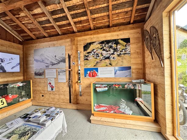 Ein gemütlicher Raum mit Holzverkleidung und Ausstellungen über Wintersport. An den Wänden hängen Bilder und Informationen, während eine Vitrine mit Ausrüstung im Vordergrund steht.