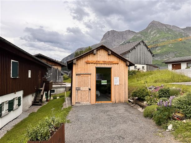 Ein Holzgebäude mit einer einladenden Eingangstür steht inmitten von grünen Wiesen und Bergen. Die Wolken am Himmel verleihen der Szene eine ruhige Atmosphäre.