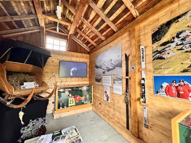 Ein gemütlicher Raum mit Holzvertäfelung, der verschiedene Ausstellungsstücke und Informationen über den Wintersport zeigt. An der Wand sind Fotos und eine Skiausrüstung zu sehen.