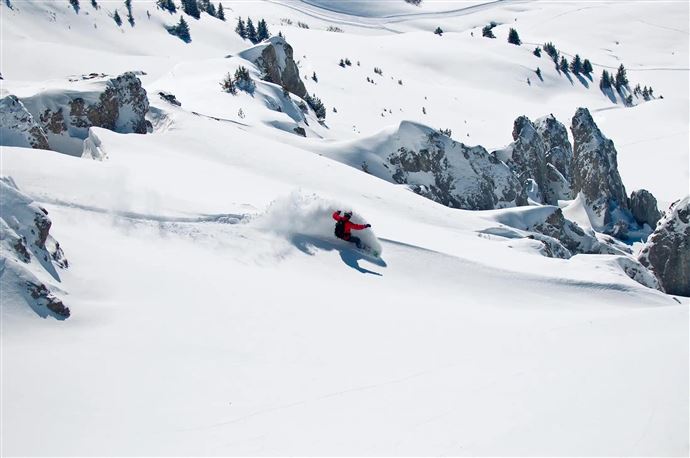 Ein Skifahrer fährt durch frischen Pulverschnee in einer beeindruckenden Berglandschaft. Die Umgebung ist von schneebedeckten Felsen und Tannen geprägt.