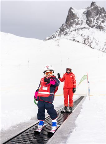A child in ski gear is standing on a snow slope and smiling. In the background, a ski instructor in red clothing can be seen.