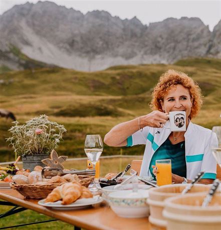 Eine Frau mit lockigem, rotem Haar sitzt an einem Tisch im Freien und genießt einen Kaffee. Der Tisch ist mit verschiedenen Speisen und Getränken auf einer malerischen Berglandschaft gedeckt.