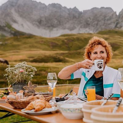 Eine Frau mit lockigem, rotem Haar sitzt an einem Tisch im Freien und genießt einen Kaffee. Der Tisch ist mit verschiedenen Speisen und Getränken auf einer malerischen Berglandschaft gedeckt.