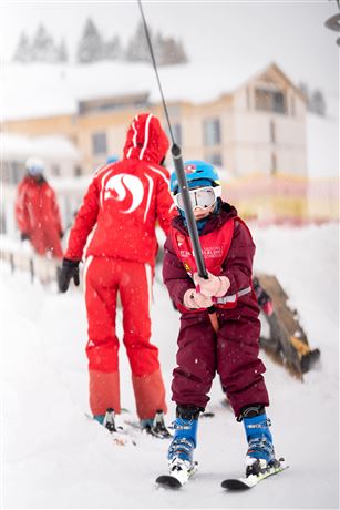 A child in a ski suit is being pulled by a ski lift, while a ski instructor in red clothing stands behind him. It is snowing and the surroundings are wintery.