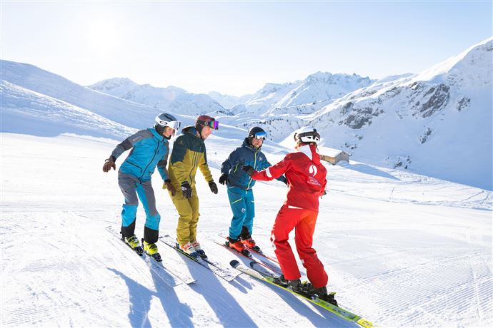 A group of skiers is standing on a snowy slope in the mountains. They are receiving instruction from a ski instructor in red gear.