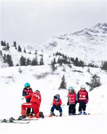 A ski instructor helps children ski in a snowy landscape. In the background, trees and mountains are visible.